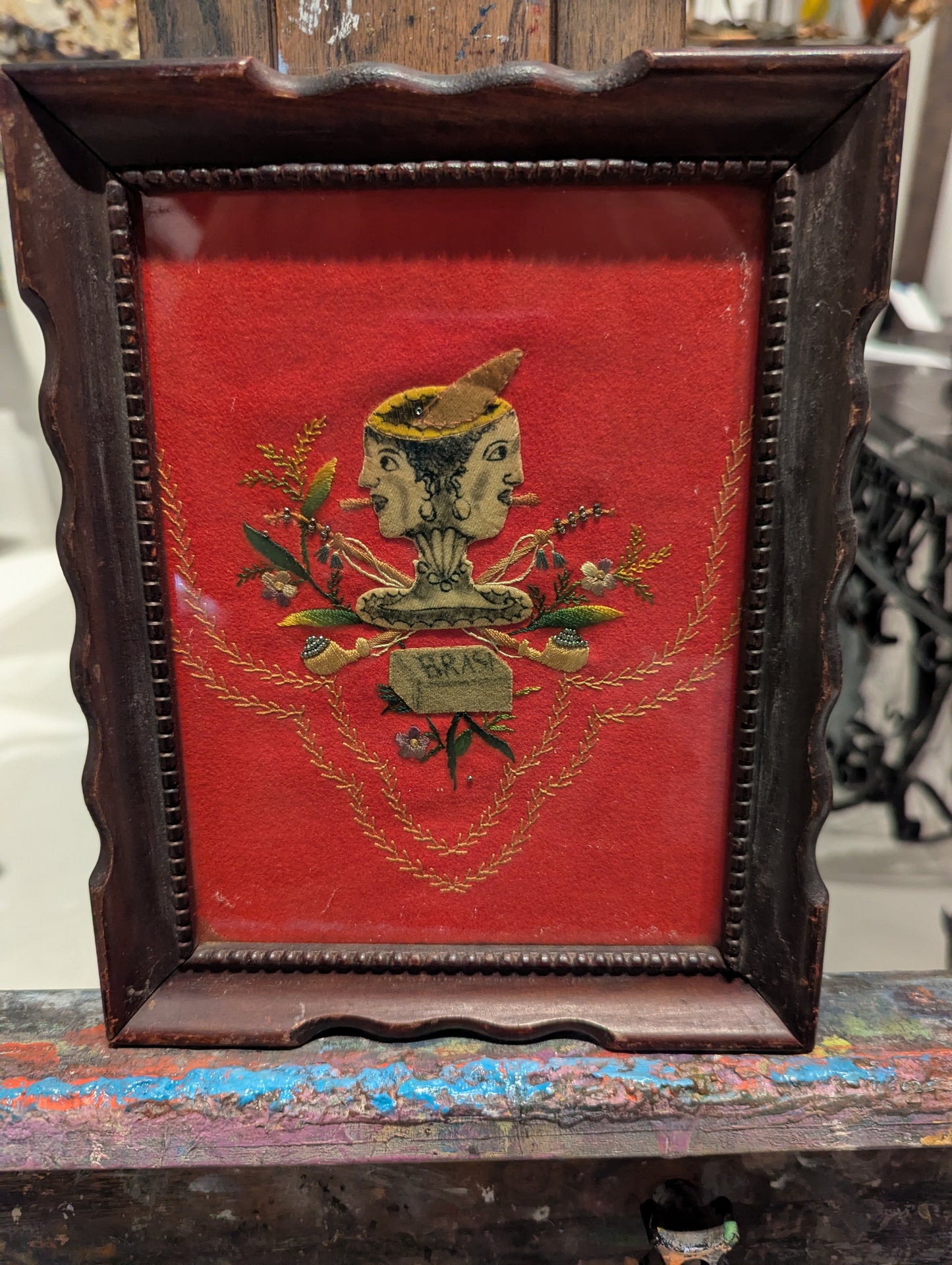 Folk art two-faced head textile on red velvet in dark scalloped frame, on easel