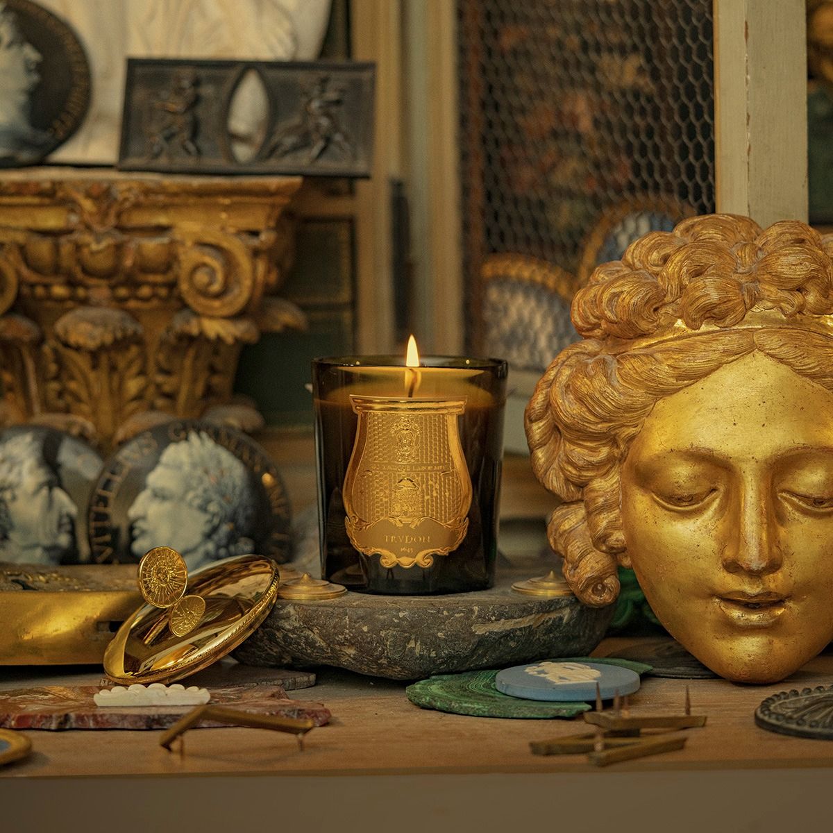 Lit Trudon Cyrnos candle styled among gilded antiques and a giltwood classical face mask.