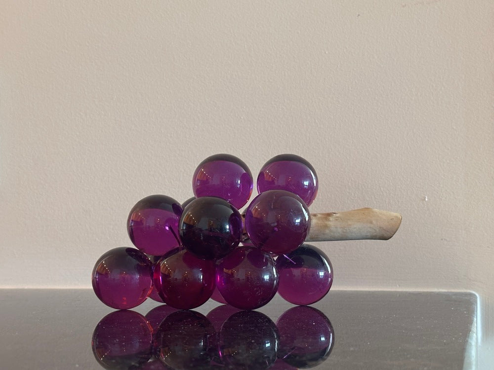 MCM purple acrylic grapes on a driftwood stem, side view on a reflective surface against a wall