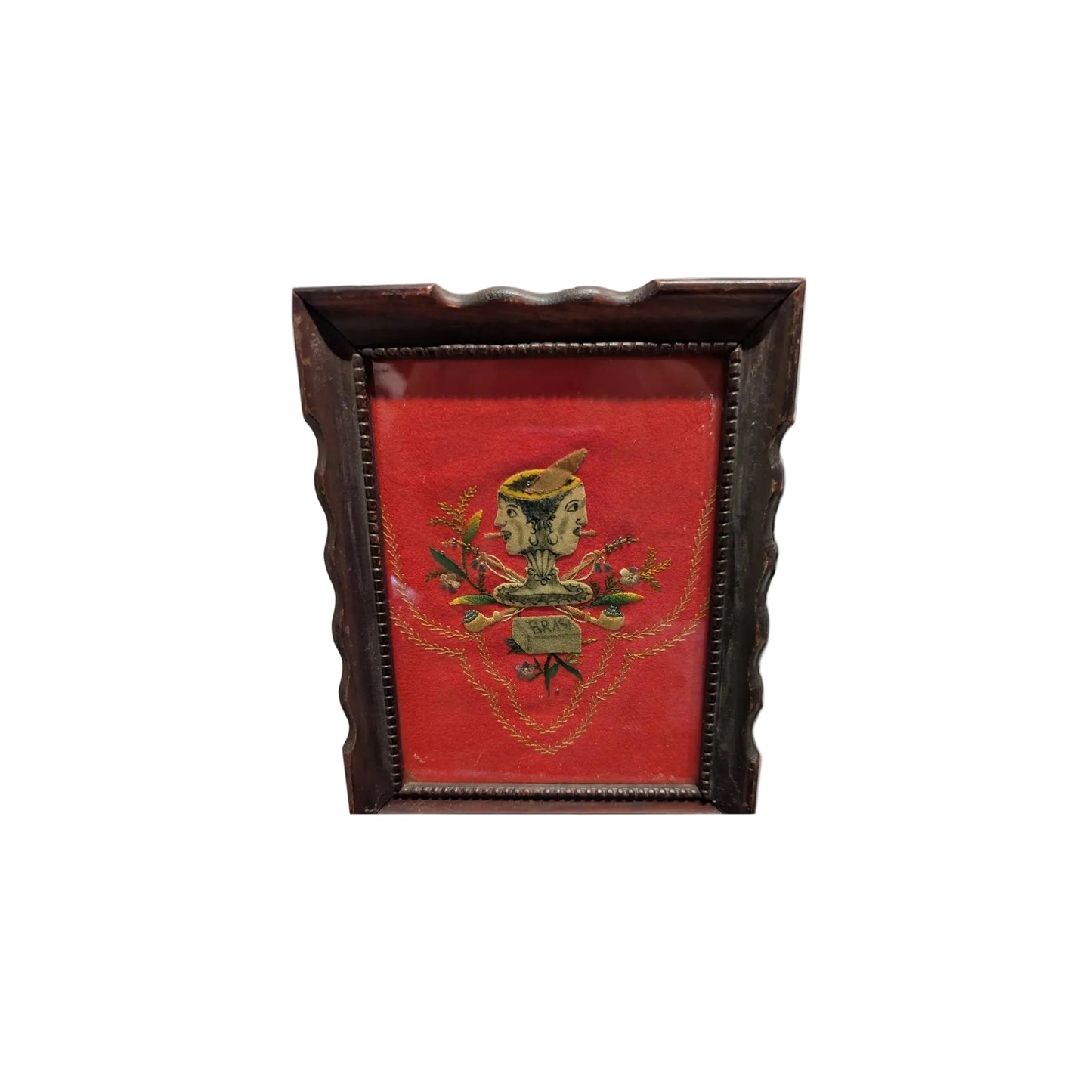 Framed folk art textile with a double-faced figure on red velvet, dark wood frame