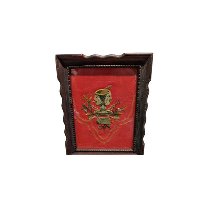 Framed folk art textile with a double-faced figure on red velvet, dark wood frame