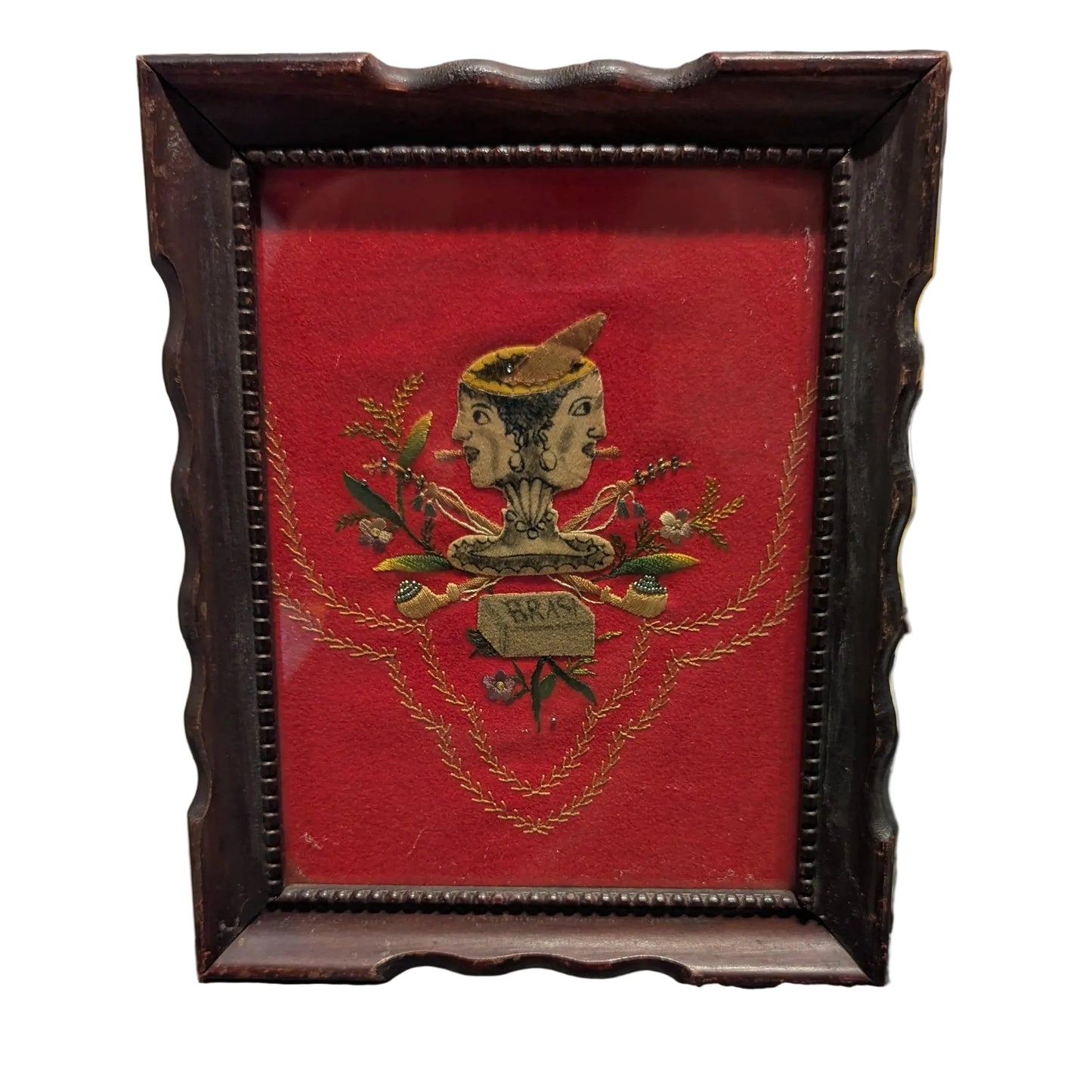 Folk art textile of a two-faced head over crossed pipes on red velvet, dark scalloped frame