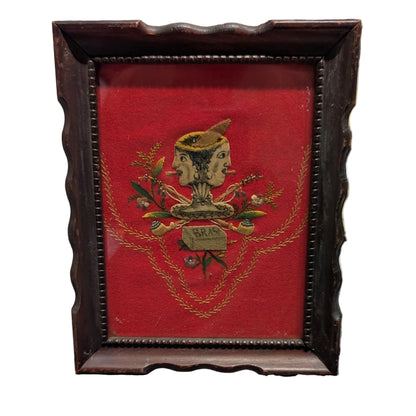 Folk art textile of a two-faced head over crossed pipes on red velvet, dark scalloped frame