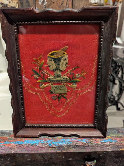 Folk art two-faced head textile on red velvet in dark scalloped frame, on easel