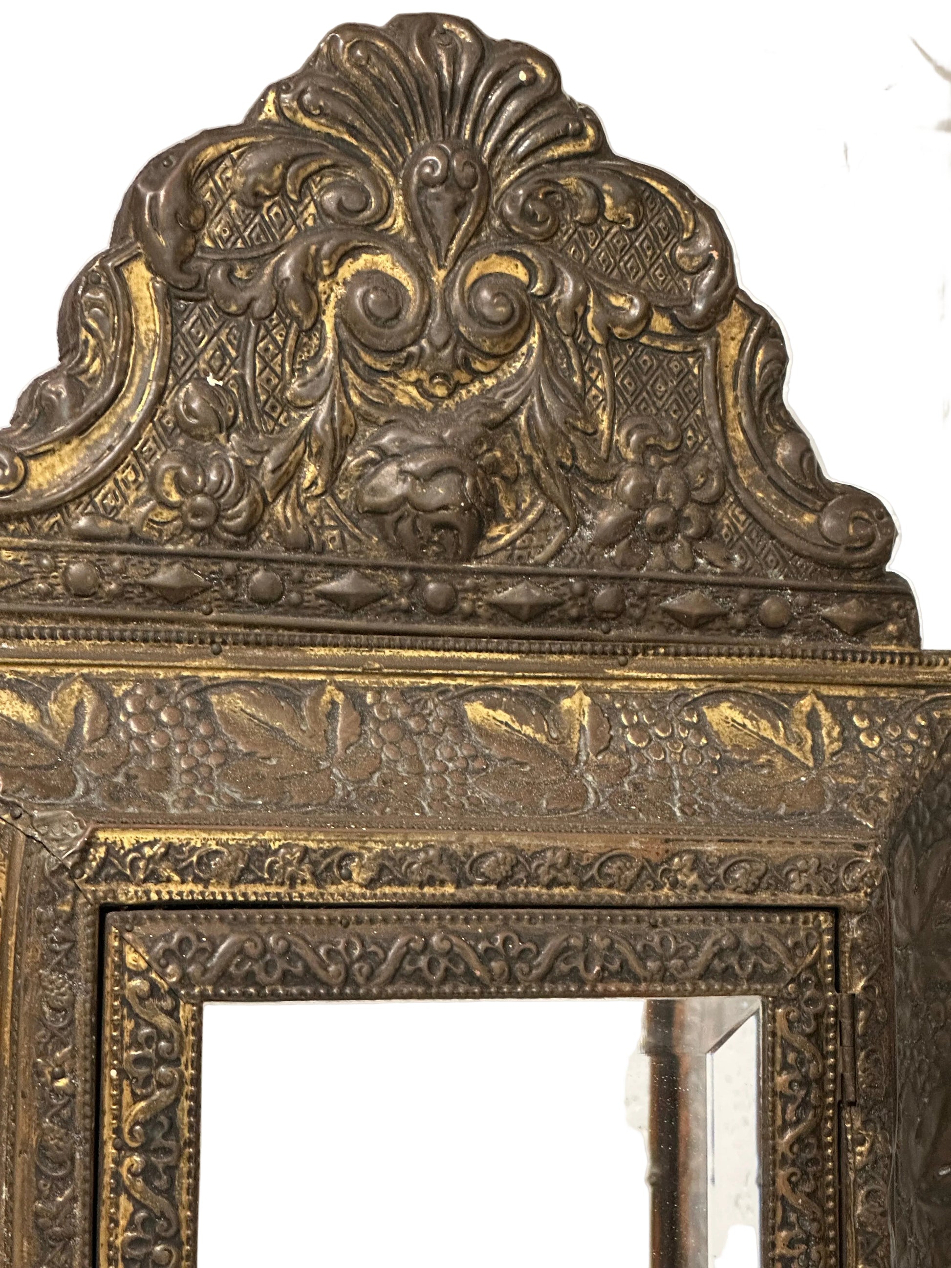 Antique embossed brass wall cabinet, top showing ornate repousse crest above mirrored door.