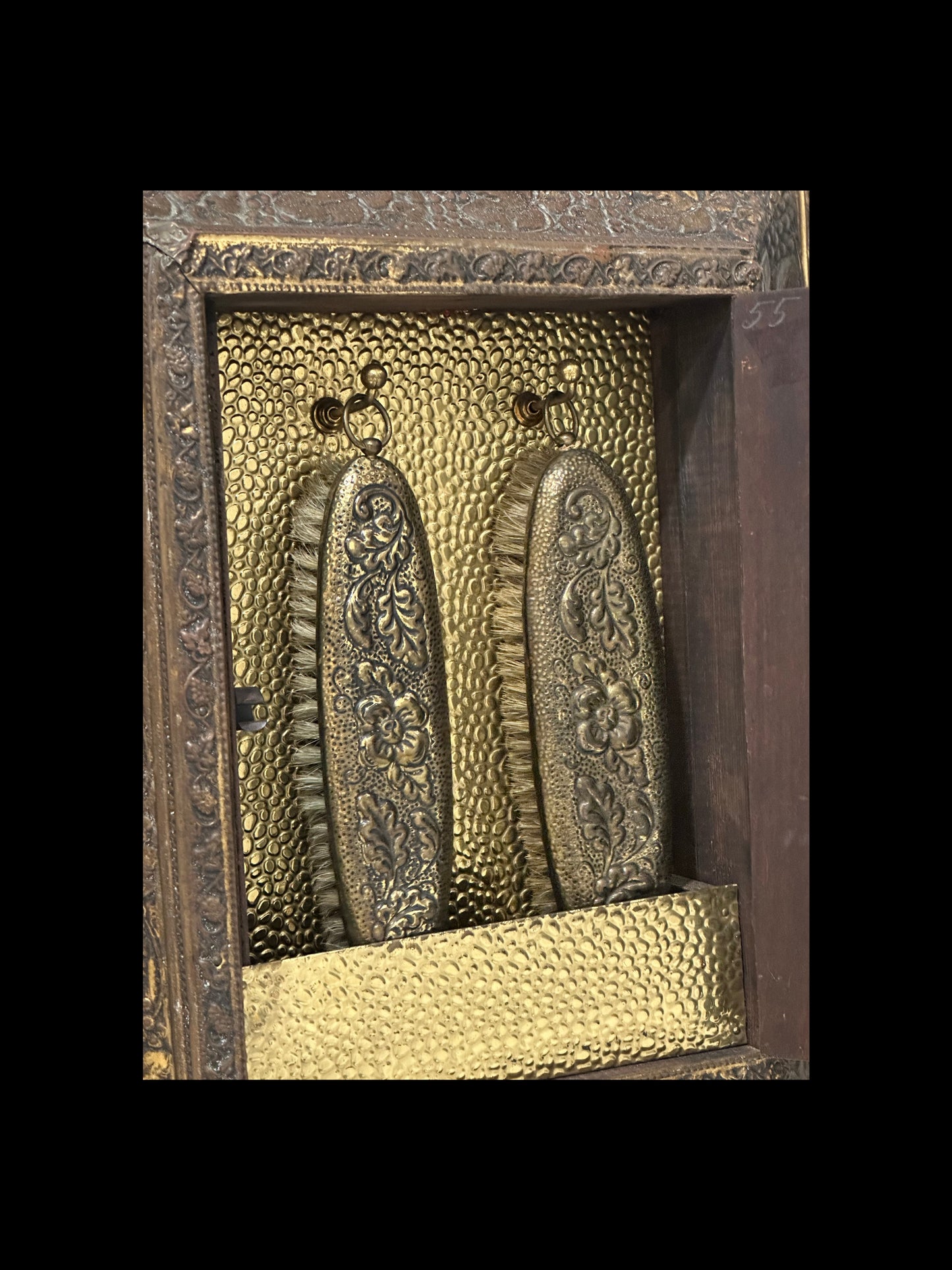 Interior of antique brass clothes-care cabinet, two floral embossed brush backs hanging on hooks.