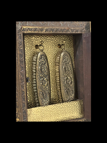 Interior of antique brass clothes-care cabinet, two floral embossed brush backs hanging on hooks.