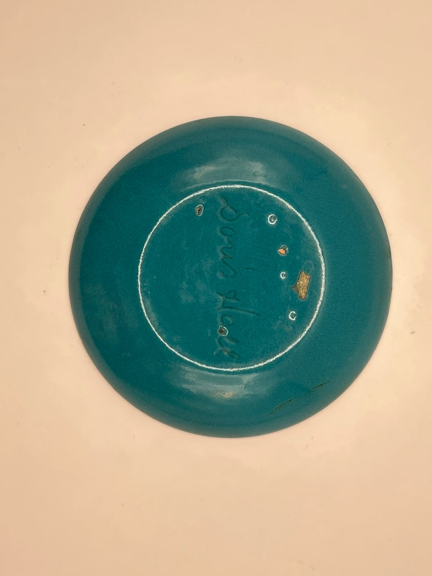 Underside of Doris Hall enamel dish on pink surface, teal base with incised signature