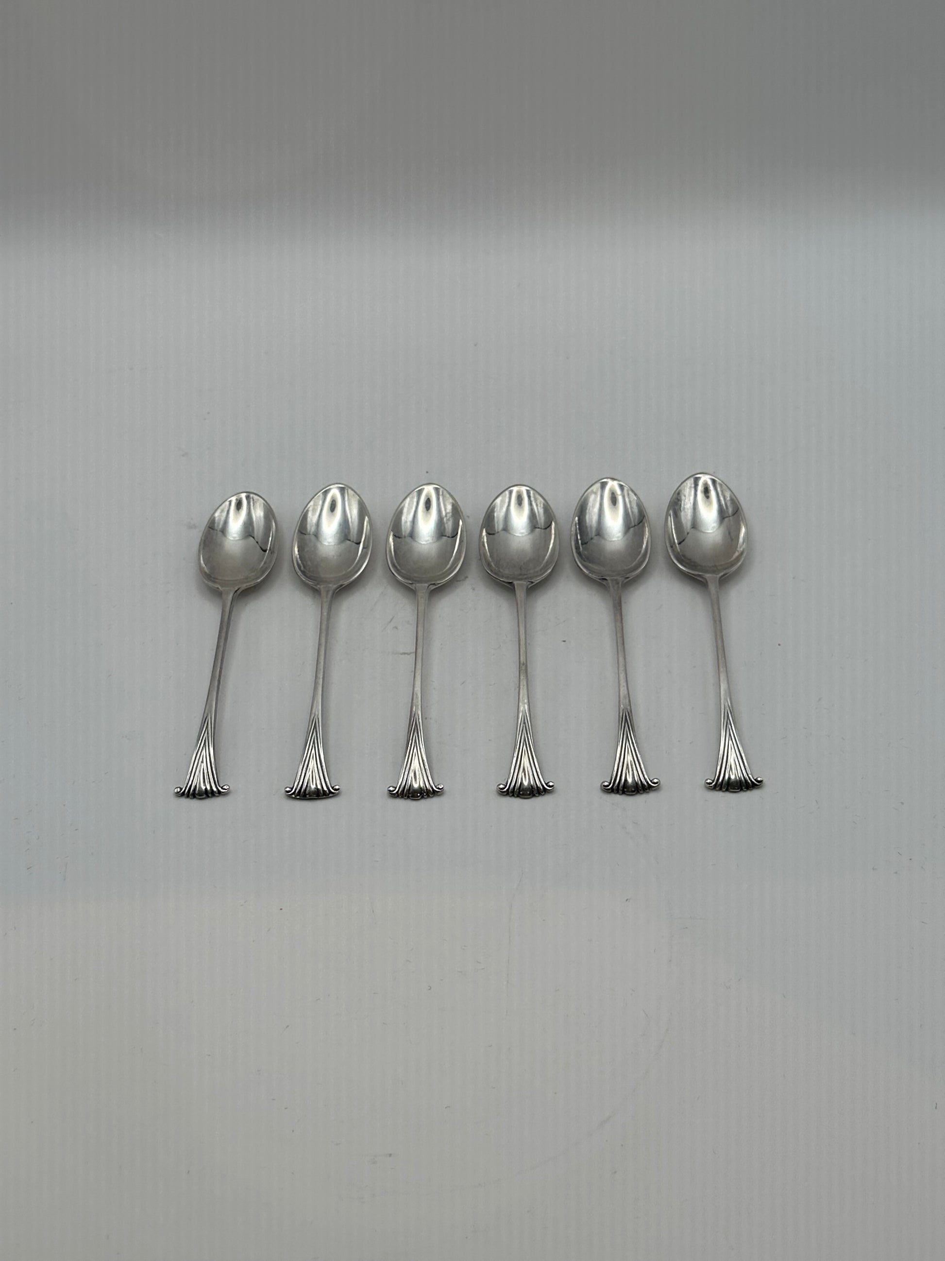 Six silverplate demitasse spoons laid in a row on a grey surface, bowls up, fanned scroll handle ends aligned
