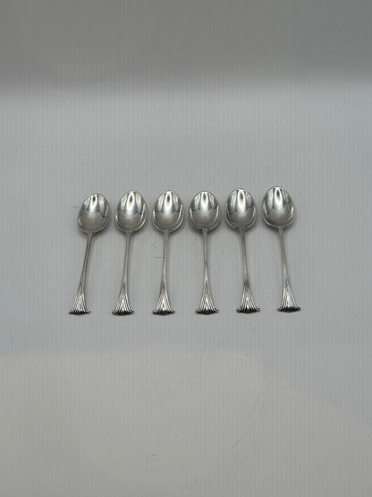 Six silverplate demitasse spoons laid in a row on a grey surface, bowls up, fanned scroll handle ends aligned