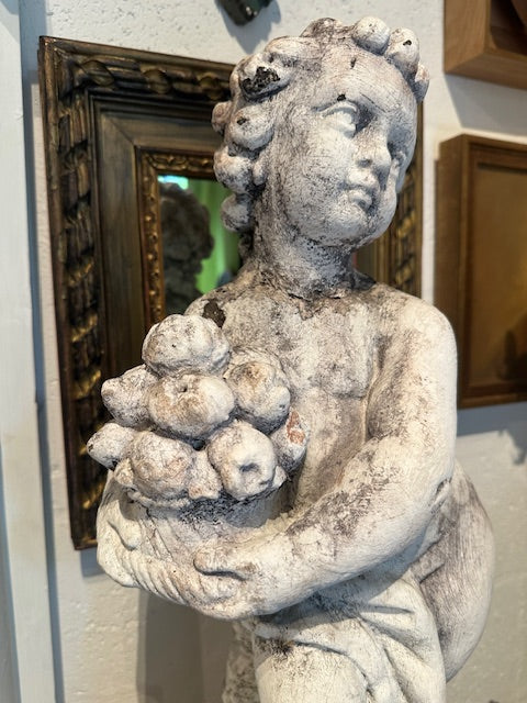 Weathered white concrete Autumn cherub statue cradling a basket of fruit, front three-quarter view