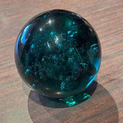 Teal controlled-bubble glass sphere on wood, angled view with shadow and clear bubbles
