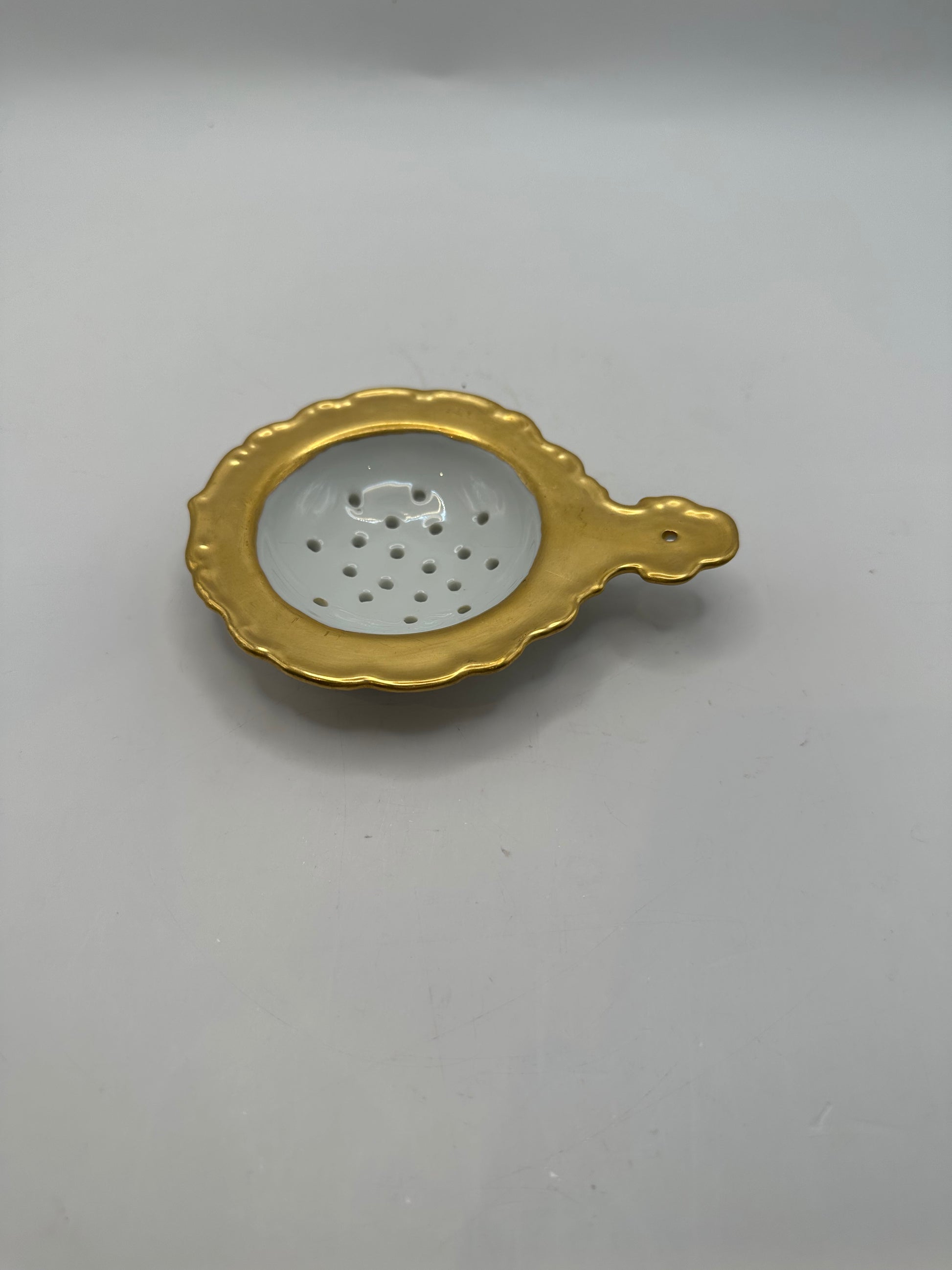 Gold-rimmed porcelain tea strainer with a scalloped edge and handle, angled view