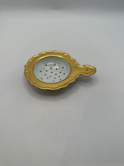 Gold-rimmed porcelain tea strainer with a scalloped edge and handle, angled view