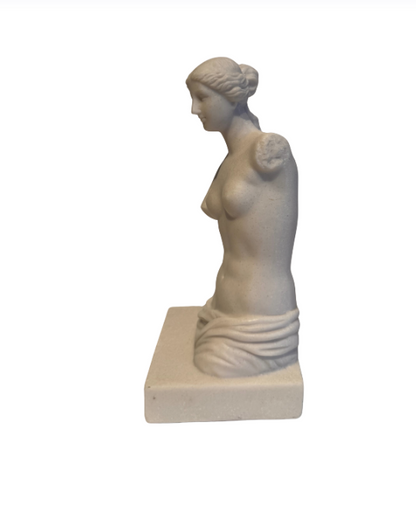 Off-white classical Venus de Milo sculpture depicting a female figure with wavy hair arranged in a bun, a softly detailed face, an armless torso, and draped fabric around the hips. The sculpture stands on a rectangular base with subtle speckling and minor surface discoloration.