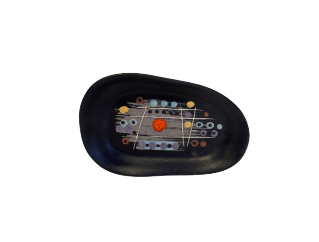 A shallow, asymmetrical oval ceramic dish with a matte black finish. The interior features a central abstract geometric design composed of gray brushstrokes, intersecting ivory lines, and circular accents in turquoise, lavender, yellow, and rust orange. A bright orange-red circle is positioned near the center. The underside is black with a handwritten "Saar" signature.