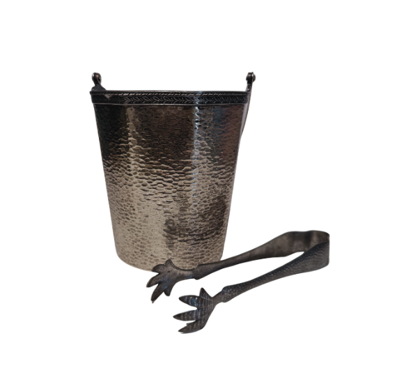 A vintage silver-tone metal ice bucket with a hexagonal, slightly tapered body and a textured hammered surface. A narrow decorative band with a repeating leaf pattern encircles the upper rim. A flat metal carrying handle is attached to opposite sides of the bucket and can be raised or lowered. The matching metal ice tongs feature textured arms ending in three-pronged bird claw shapes. The bucket's interior has a hammered texture with visible darkening and discoloration at the bottom.