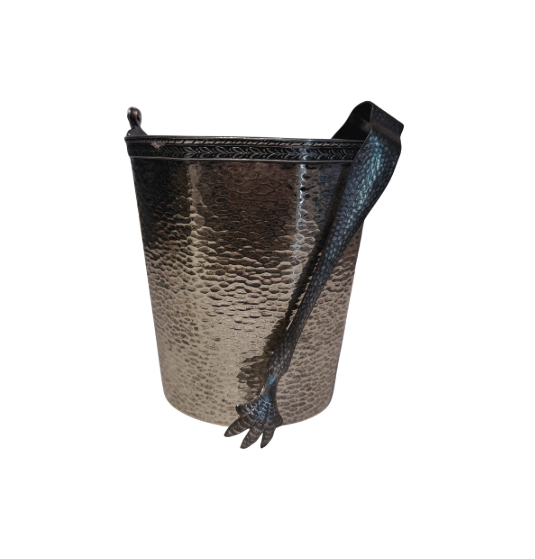 A vintage silver-tone metal ice bucket with a hexagonal, slightly tapered body and a textured hammered surface. A narrow decorative band with a repeating leaf pattern encircles the upper rim. A flat metal carrying handle is attached to opposite sides of the bucket and can be raised or lowered. The matching metal ice tongs feature textured arms ending in three-pronged bird claw shapes. The bucket's interior has a hammered texture with visible darkening and discoloration at the bottom.