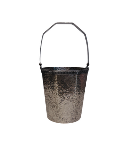 A vintage silver-tone metal ice bucket with a hexagonal, slightly tapered body and a textured hammered surface. A narrow decorative band with a repeating leaf pattern encircles the upper rim. A flat metal carrying handle is attached to opposite sides of the bucket and can be raised or lowered. The matching metal ice tongs feature textured arms ending in three-pronged bird claw shapes. The bucket's interior has a hammered texture with visible darkening and discoloration at the bottom.