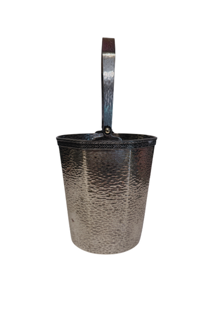 A vintage silver-tone metal ice bucket with a hexagonal, slightly tapered body and a textured hammered surface. A narrow decorative band with a repeating leaf pattern encircles the upper rim. A flat metal carrying handle is attached to opposite sides of the bucket and can be raised or lowered. The matching metal ice tongs feature textured arms ending in three-pronged bird claw shapes. The bucket's interior has a hammered texture with visible darkening and discoloration at the bottom.