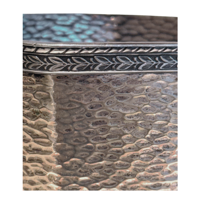 A vintage silver-tone metal ice bucket with a hexagonal, slightly tapered body and a textured hammered surface. A narrow decorative band with a repeating leaf pattern encircles the upper rim. A flat metal carrying handle is attached to opposite sides of the bucket and can be raised or lowered. The matching metal ice tongs feature textured arms ending in three-pronged bird claw shapes. The bucket's interior has a hammered texture with visible darkening and discoloration at the bottom.