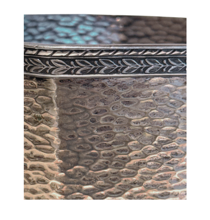 A vintage silver-tone metal ice bucket with a hexagonal, slightly tapered body and a textured hammered surface. A narrow decorative band with a repeating leaf pattern encircles the upper rim. A flat metal carrying handle is attached to opposite sides of the bucket and can be raised or lowered. The matching metal ice tongs feature textured arms ending in three-pronged bird claw shapes. The bucket's interior has a hammered texture with visible darkening and discoloration at the bottom.