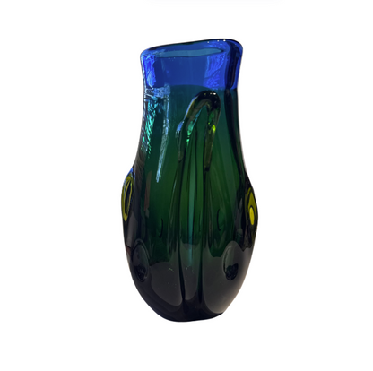 Tall, sculptural glass vase with a deep emerald green body, rounded base, and thick cobalt blue rim. Raised, flowing glass accents extend along the sides, incorporating translucent yellow-green details. The vase has an irregular oval opening and a glossy, reflective surface.