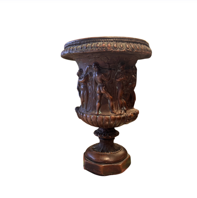 Ornate Neoclassical-style decorative urn with a dark bronze finish, raised classical figures, scrolling foliage, sculptural masks, a wide patterned rim, and a fluted pedestal base. Measures 6.5 inches in diameter and 9.25 inches tall.