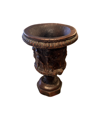 Ornate Neoclassical-style decorative urn with a dark bronze finish, raised classical figures, scrolling foliage, sculptural masks, a wide patterned rim, and a fluted pedestal base. Measures 6.5 inches in diameter and 9.25 inches tall.