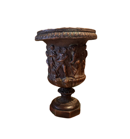 Ornate Neoclassical-style decorative urn with a dark bronze finish, raised classical figures, scrolling foliage, sculptural masks, a wide patterned rim, and a fluted pedestal base. 