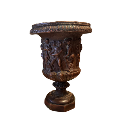 Ornate Neoclassical-style decorative urn with a dark bronze finish, raised classical figures, scrolling foliage, sculptural masks, a wide patterned rim, and a fluted pedestal base. 