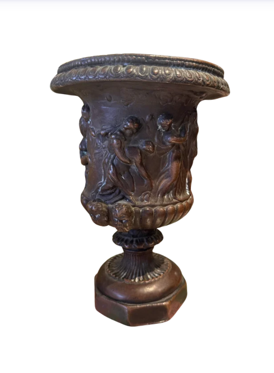 Ornate Neoclassical-style decorative urn with a dark bronze finish, raised classical figures, scrolling foliage, sculptural masks, a wide patterned rim, and a fluted pedestal base. 