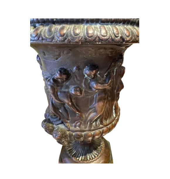 Ornate Neoclassical-style decorative urn with a dark bronze finish, raised classical figures, scrolling foliage, sculptural masks, a wide patterned rim, and a fluted pedestal base.  Close up 