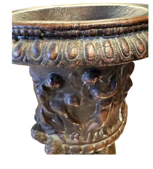 Ornate Neoclassical-style decorative urn with a dark bronze finish, raised classical figures, scrolling foliage, sculptural masks, a wide patterned rim, and a fluted pedestal base. Close up of rim
