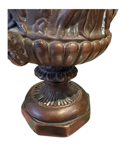 Ornate Neoclassical-style decorative urn with a dark bronze finish, raised classical figures, scrolling foliage, sculptural masks, a wide patterned rim, and a fluted pedestal base. Close up of base