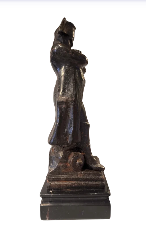 Side View of Dark cast iron statue depicting Napoleon Bonaparte standing with his arms crossed, wearing a bicorne military hat, long overcoat, and boots. The figure stands on a stepped rectangular black marble base.