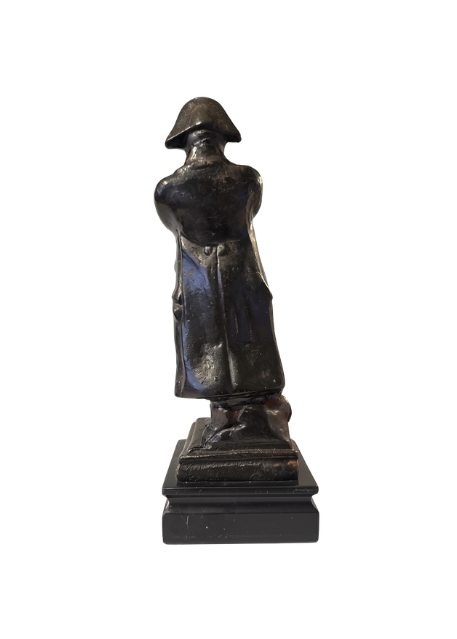 back View of Dark cast iron statue depicting Napoleon Bonaparte standing with his arms crossed, wearing a bicorne military hat, long overcoat, and boots. The figure stands on a stepped rectangular black marble base.