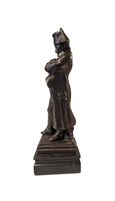 Side View of Dark cast iron statue depicting Napoleon Bonaparte standing with his arms crossed, wearing a bicorne military hat, long overcoat, and boots. The figure stands on a stepped rectangular black marble base.