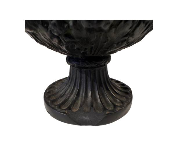 Close up View of the foot of the urn with the chipped edge, Black faux-bronze Neoclassical Medici urn with green patina accents, measuring 11 inches tall and 9 inches in diameter. The Campana-shaped vessel features a wide flared rim with egg-and-dart detailing, raised Greco-Roman figures in various poses, grapevine and leaf borders, and two reeded loop handles with bearded mask decorations. An intricate floral and acanthus leaf design surrounds the lower bowl, which rests on a fluted circular pedestal base.