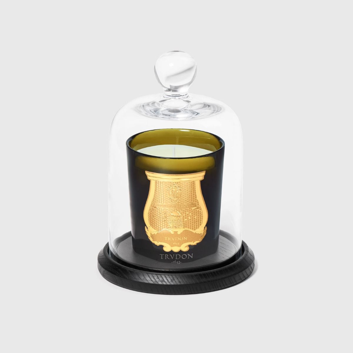 Trudon green candle with gold crest under a clear glass cloche on a black wood base
